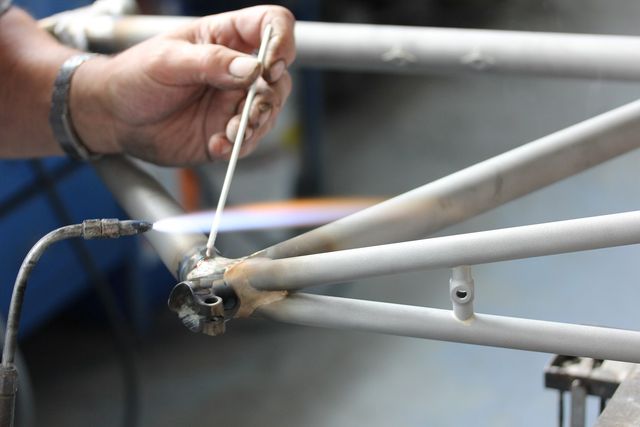 自転車フレームの鋼管のつなぎ目を炎で加熱し棒状のろうを当てている
